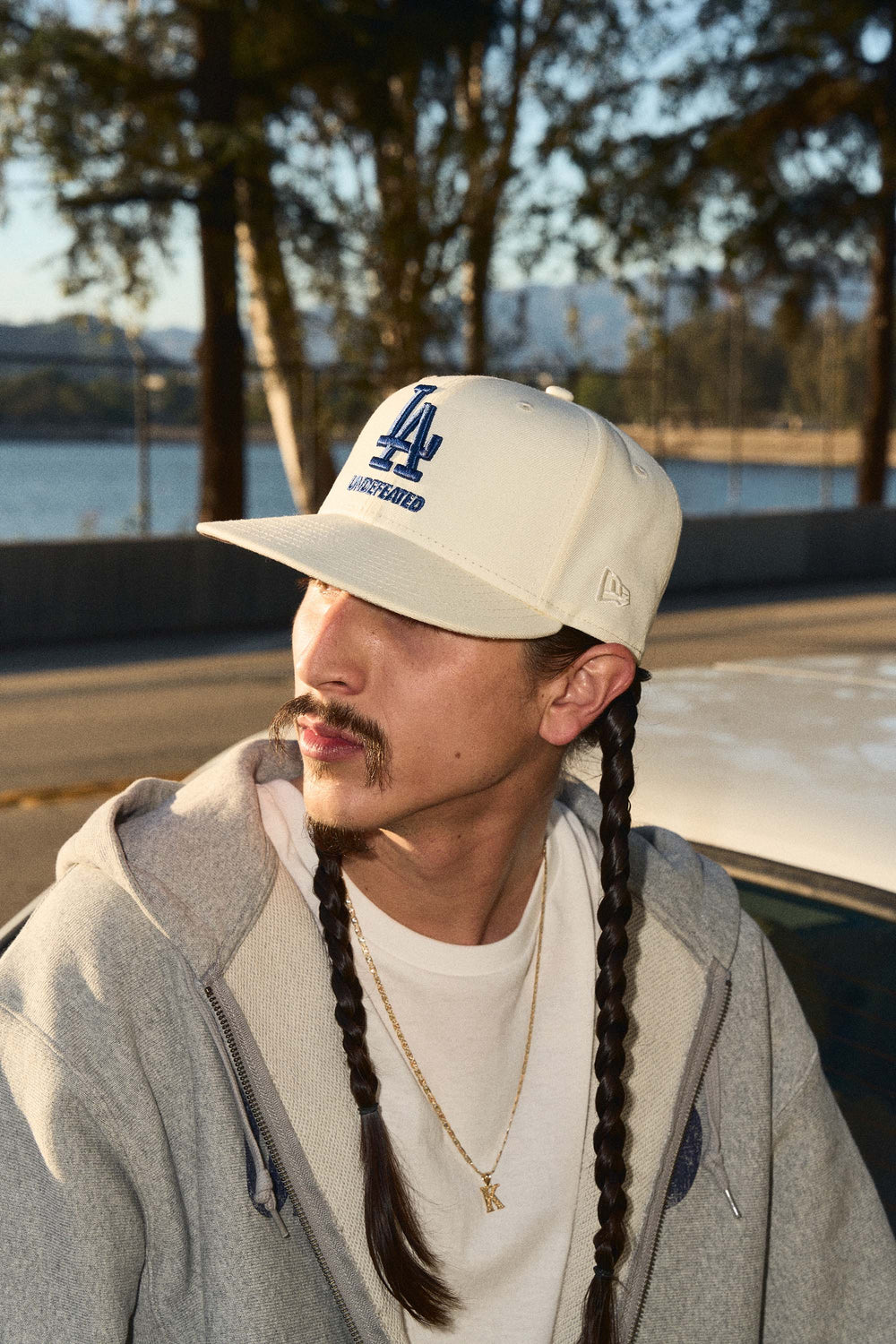 Person wearing a white cap with a logo, gray hoodie, and white shirt outdoors by a body of water.